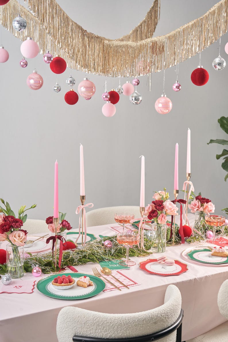Puede incluir: Un mantel rosa con un plato estampado en verde y blanco, un plato estampado en rosa y rojo y una copa de champ&aacute;n rosado. Hay velas, flores y una guirnalda de oropel dorado y adornos rojos y rosas colgando sobre la mesa.