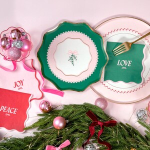 Puede incluir: Platos de papel verde y rosa con borde dorado y centro blanco con una rama de acebo verde con un lazo rosa. Los platos est&aacute;n colocados sobre una superficie rosa con servilletas verdes y rosas que dicen "JOY", "PEACE" y "LOVE".