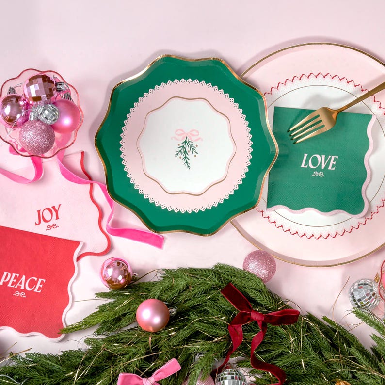 Puede incluir: Platos de papel verde y rosa con borde dorado y centro blanco con una rama de acebo verde con un lazo rosa. Los platos est&aacute;n colocados sobre una superficie rosa con servilletas verdes y rosas que dicen "JOY", "PEACE" y "LOVE".