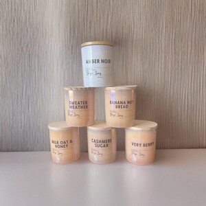Könnte beinhalten: Sechs Duftkerzen mit Holzdeckeln. Die Kerzen sind in verschiedenen Farben, darunter Weiß, Rosa und Orange. Die Kerzenaufkleber tragen die Namen "Amber Noir", "Sweater Weather", "Banana Nut Bread", "Milk Oat & Honey", "Cashmere Sugar" und "Very Berry".
