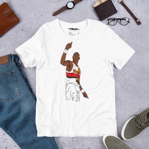 Può includere: Maglietta bianca con una grafica di un giocatore di basket che indossa una maglia rossa, bianca e nera con la scritta "Bulls" sul davanti. Il giocatore sta alzando la mano destra in aria e puntando con la mano sinistra.