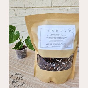 May include: A brown paper bag with a clear window showing a mix of potting soil for aroids. The bag has a label that reads "Aroid Mix" and lists the ingredients: pine bark, coco chips, coco coir, chunky perlite, leca, horticultural charcoal, and worm castings. The label also states that the mix is perfect for aroids such as philodendron, monstera, alocasia, anthurium, pothos, syngonium, etc.