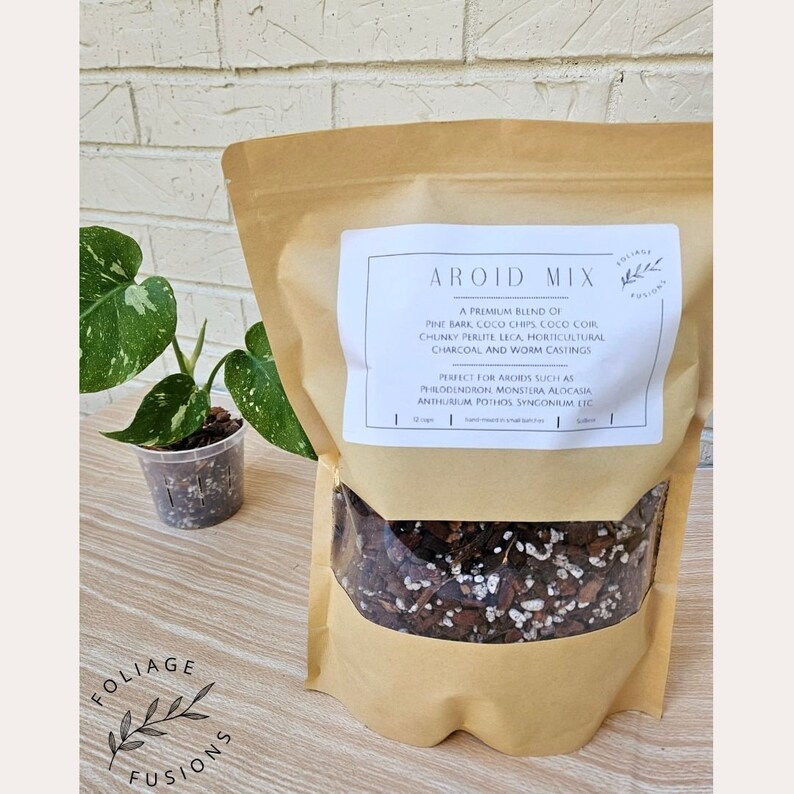 May include: A brown paper bag with a clear window showing a mix of potting soil for aroids. The bag has a label that reads "Aroid Mix" and lists the ingredients: pine bark, coco chips, coco coir, chunky perlite, leca, horticultural charcoal, and worm castings. The label also states that the mix is perfect for aroids such as philodendron, monstera, alocasia, anthurium, pothos, syngonium, etc.