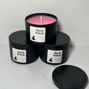 May include: Three black candle jars with lids. The jars are labeled "Basic Witch" with a witch hat graphic. The candle in the front jar is pink.