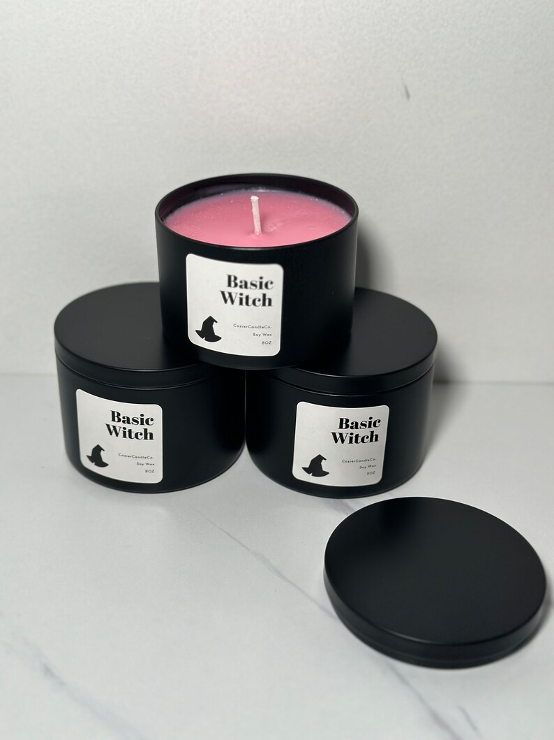 May include: Three black candle jars with lids. The jars are labeled "Basic Witch" with a witch hat graphic. The candle in the front jar is pink.