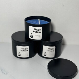 以下が含まれることがあります： 青いワックスと蓋が付いた黒いキャンドルジャーが4つ。ジャーには、「マジックポーション」という文字と、ポーションボトルの小さなイラストが描かれた白いラベルが付いています。