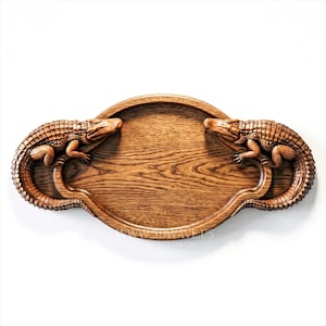 May include: Wooden serving tray with two detailed alligator sculptures on either side. The tray is oval-shaped with a light brown wood grain finish. The alligators are a darker brown color.