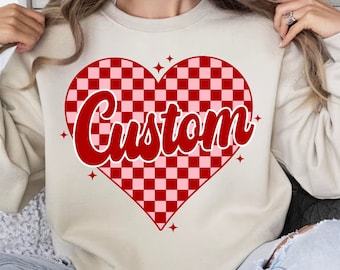 May include: A cream-coloured sweatshirt with a red and pink checkered heart design. The heart has the word "Custom" written in a red, retro font.