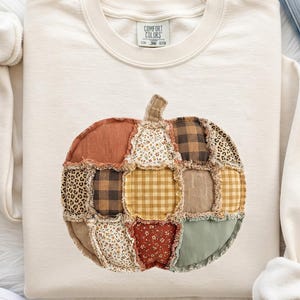 May include: A cream-colored crewneck sweatshirt featuring a central pumpkin graphic made of various fabric patches with raw, frayed edges. The patches include patterns like leopard print, brown and black buffalo check, yellow gingham, and floral prints.