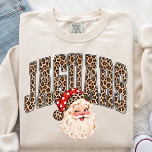 May include: A cream-colored crewneck sweatshirt featuring the word JAGUARS in a leopard print pattern with a black outline. Below the text is a graphic of a winking Santa Claus wearing a red and white polka dot hat with leopard print trim.