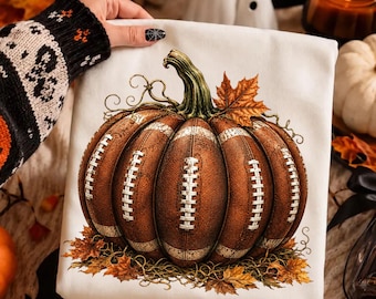 May include: An ivory sweatshirt featuring a graphic of a pumpkin designed with the texture and white laces of a brown football. The pumpkin is surrounded by orange and brown autumn leaves and thin, curling vines, creating a seasonal aesthetic.