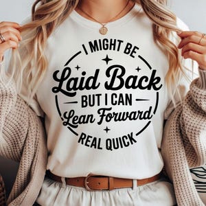 May include: A cream-coloured t-shirt featuring the text "I MIGHT BE Laid Back BUT I CAN Lean Forward REAL QUICK" in a black, circular design. The shirt has a crew neck and is worn with a brown belt and beige trousers.