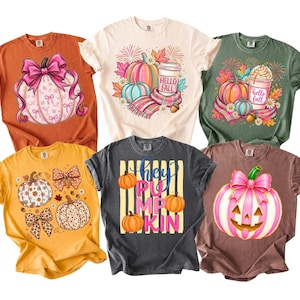 Pode incluir: Uma coleção de seis camisetas de manga curta em várias cores com gráficos de temática de outono. Os designs incluem abóboras com laços cor-de-rosa, copos de café com a etiqueta Hello Fall, abóboras com laços de leopardo e o texto hey PUMPKIN.