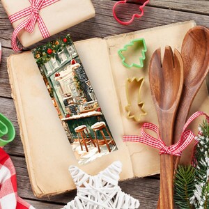 May include: A Christmas-themed bookmark with a colorful illustration of a storefront with snow falling. The bookmark is laying open on a wooden table with a cookbook, wooden spoons, and a Christmas tree branch.