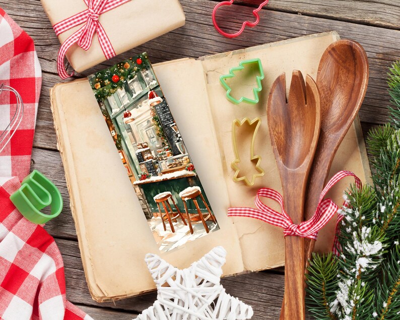 May include: A Christmas-themed bookmark with a colorful illustration of a storefront with snow falling. The bookmark is laying open on a wooden table with a cookbook, wooden spoons, and a Christmas tree branch.