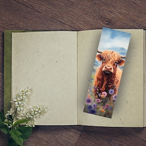 May include: A bookmark featuring a close-up of a Highland cow with long, reddish-brown fur standing in a field of purple and pink wildflowers. The background is a blurred image of a mountain range and a blue sky.