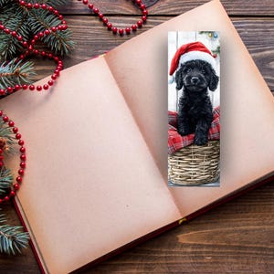 May include: A bookmark featuring a black puppy wearing a Santa hat, sitting in a woven basket lined with red plaid fabric. The bookmark is set against a wooden background, with an open book and Christmas decorations.