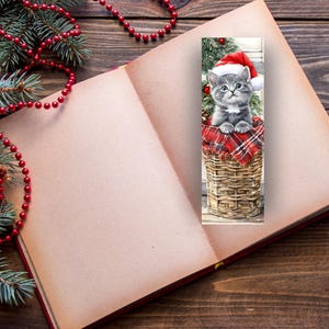 May include: A Christmas-themed bookmark featuring a gray kitten wearing a Santa hat, sitting in a woven basket lined with red plaid fabric. The bookmark is placed in an open book, with Christmas decorations in the background.