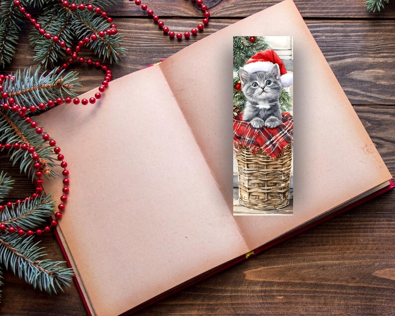 May include: A Christmas-themed bookmark featuring a gray kitten wearing a Santa hat, sitting in a woven basket lined with red plaid fabric. The bookmark is placed in an open book, with Christmas decorations in the background.