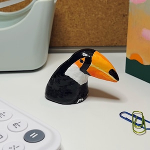 Peut inclure: Une figurine de toucan faite à la main, avec un corps noir, un visage blanc et un grand bec orange et jaune. Le toucan est sur un bureau blanc avec une calculatrice, un distributeur de ruban adhésif et un porte-stylos peint. Deux trombones sont également sur le bureau.