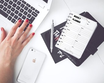 May include: A white weekly planner with a spiral binding, open to a blank page, sits atop a black leather-bound notebook. A laptop, pen, and smartphone are also on the white surface. The planner has the words "WEEKLY PLANNER" at the top.