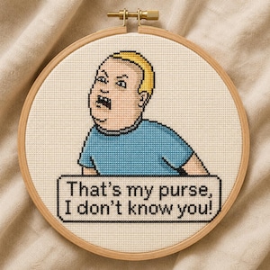 May include: A circular wooden embroidery hoop containing a cross-stitch design of a cartoon character with blonde hair and a blue shirt. Below the character, a rectangular speech bubble contains the black text: That's my purse, I don't know you!