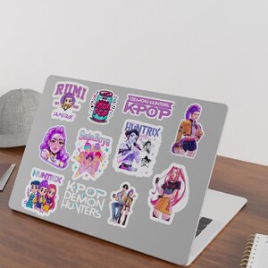 May include: A laptop covered in colorful stickers featuring anime-style illustrations and text. The stickers showcase various characters and phrases, including "RUMI," "KPOP," and "DEMON HUNTERS." The laptop sits on a wooden desk with a small potted plant and a notebook.