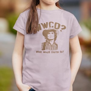 May include: A light pink t-shirt with a brown graphic of a man wearing a cowboy hat. The graphic says "WWCD? What would Charles do?"