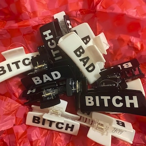 May include: A collection of black and white hair claw clips. The clips are rectangular with the words "BAD" and "BITCH" printed in bold, white letters. The clips are scattered on a pink and red checkered tissue paper.