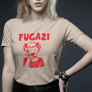 May include: A beige t-shirt with a red graphic of a cartoon girl on the phone and the text "FUGAZI" above her.