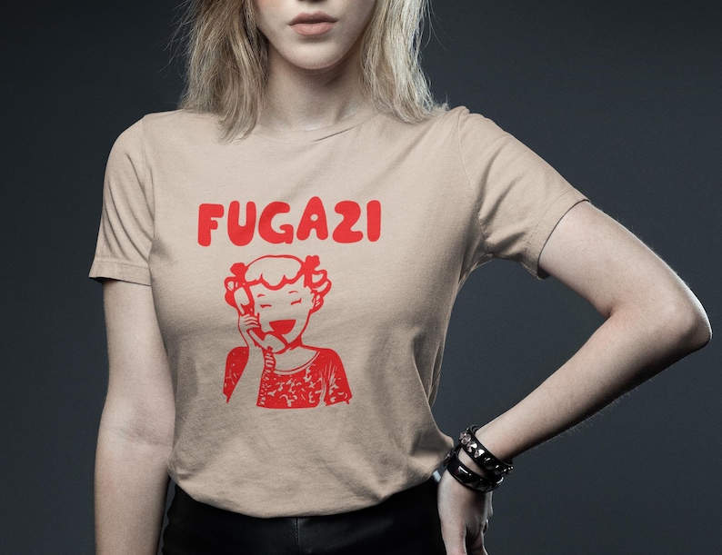 May include: A beige t-shirt with a red graphic of a cartoon girl on the phone and the text "FUGAZI" above her.
