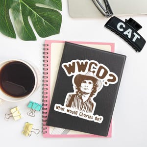 May include: A black notebook with a sticker featuring a sepia-toned image of a man in a cowboy hat, with the text "WWCD? What would Charles do?" A cup of coffee, a pink notebook, and a black hair clip are also visible.