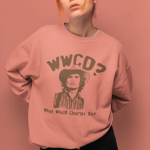 May include: A pink sweatshirt with a brown graphic print of a man wearing a cowboy hat. The text "WWCD?" and "What would Charles do?" are printed on the sweatshirt.
