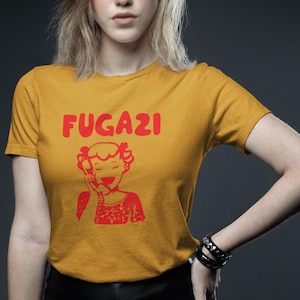 May include: A mustard yellow t-shirt with a red graphic of a cartoon girl on the phone and the word "FUGAZI" in red letters.