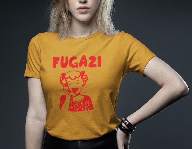 May include: A mustard yellow t-shirt with a red graphic of a cartoon girl on the phone and the word "FUGAZI" in red letters.