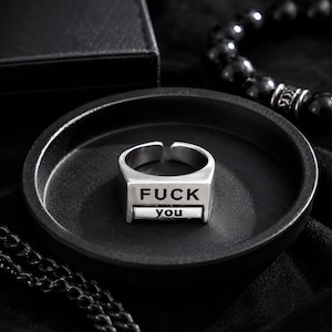 May include: A silver-toned metal ring featuring a rectangular face with the text FUCK engraved in large capital letters and the word you engraved in smaller lowercase letters on a separate horizontal bar positioned directly below the first line.