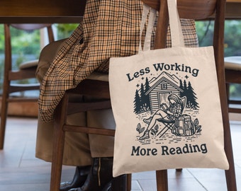 Könnte beinhalten: Eine weiße Canvas-Tragetasche mit schwarzem Text, der "Weniger Arbeiten, mehr Lesen" lautet. Die Tasche zeigt eine Schwarzweißillustration einer Person, die auf einem Stuhl sitzt und ein Buch vor einer Hütte im Wald liest.