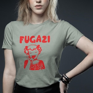 May include: A light green t-shirt with the word "FUGAZI" printed in red letters. A cartoon image of a girl on the phone is printed below the text.