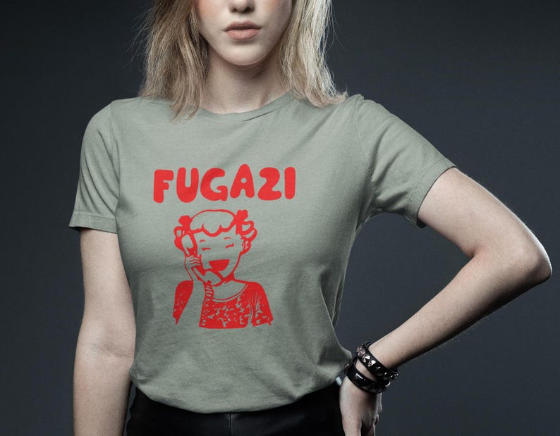 May include: A light green t-shirt with the word "FUGAZI" printed in red letters. A cartoon image of a girl on the phone is printed below the text.