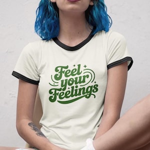 Puede incluir: Una camiseta blanca con mangas de color negro y un gráfico verde que dice "Feel your Feelings" en una fuente retro.