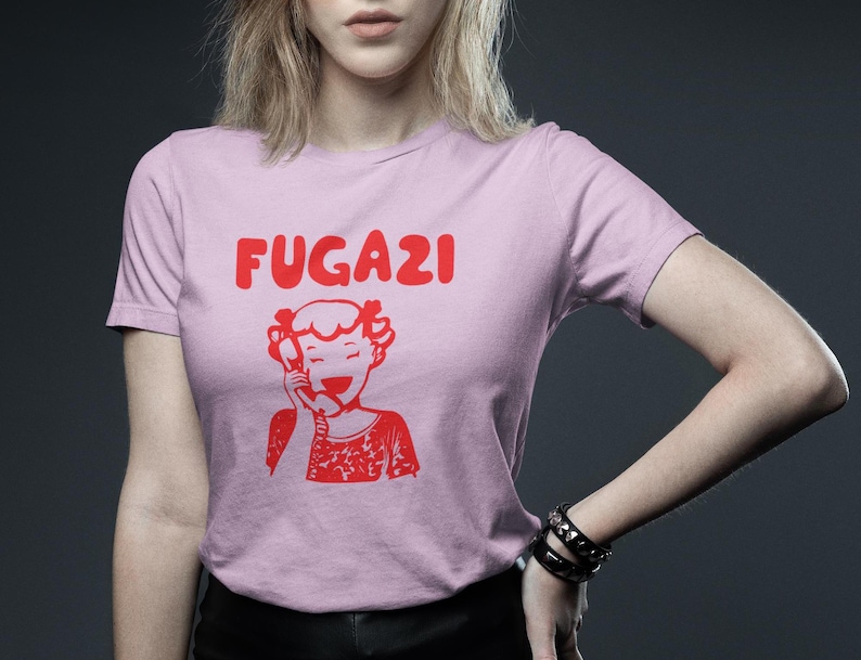 May include: Pink t-shirt with a red graphic of a girl on the phone and the word "FUGAZI" above her.