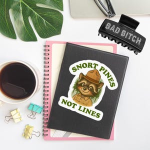May include: A black hair clip with the text "BAD BITCH" in white lettering. A black notebook with a sticker that says "SNORT PINES NOT LINES" featuring a raccoon wearing a ranger hat. A cup of coffee and office supplies are also visible.
