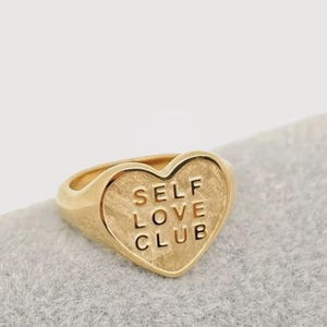 May include: A gold-colored ring with a heart-shaped face. The face of the ring has the words "SELF LOVE CLUB" engraved in capital letters. The ring is set on a light gray surface.