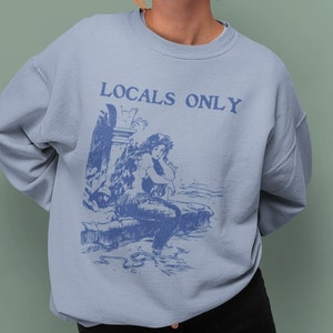 May include: Light blue sweatshirt with a blue illustration of a mermaid and the text "LOCALS ONLY" printed on the front.