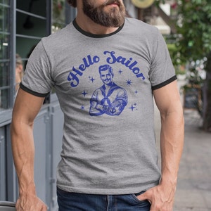 May include: A gray t-shirt with a black ringer, featuring a vintage illustration of a sailor with the text "Hello Sailor" in a blue script font.