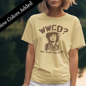 Peut inclure: Un t-shirt jaune clair avec un motif brun d'un homme portant un chapeau de cowboy et le texte "WWCD? What would Charles do?"