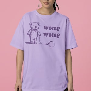 Può includere: Una maglietta color lavanda con un orsacchiotto di cartone animato che tiene un palloncino. Il testo "womp womp" è stampato sulla maglietta.