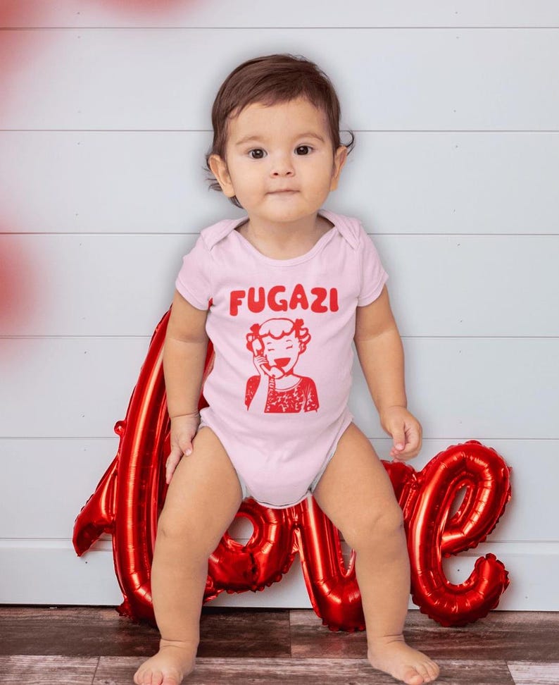 May include: A baby wearing a pink bodysuit with the word "FUGAZI" and a red cartoon image of a woman on the phone. The baby is sitting in front of a red foil balloon that says "LOVE".