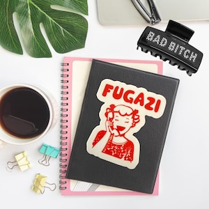 May include: A black hair claw clip with the text "BAD BITCH" in white letters. A black notebook with a sticker that says "FUGAZI" in red letters. A cup of coffee, binder clips, and a green leaf are also visible.