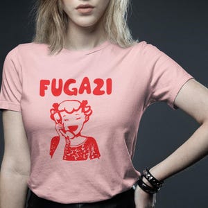 May include: A pink t-shirt with a red graphic of a girl on the phone and the word "FUGAZI" above her.